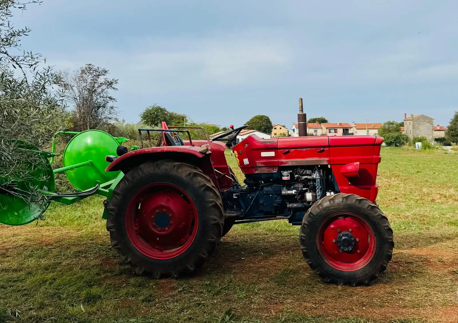 Ein roter Traktor mit grüner Netzrolle steht auf einer Wiese, im Hintergrund die Häuser eines Dorfes.